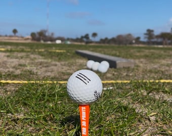 Può includere: Una palla da golf bianca con tre linee nere su di essa si trova su un tee con il testo "PAR 38 PRINTS" su di esso. La palla è su una superficie erbosa verde con un cielo blu sullo sfondo.