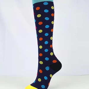May include: A knee-high sock with a dark blue base featuring a repeating pattern of orange, light blue, and lime green polka dots. The sock has a solid light blue cuff at the top, a solid black heel section, and a solid yellow toe cap.