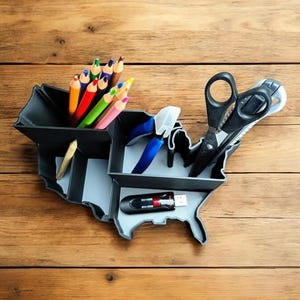 May include: A grey desk organiser shaped like the United States of America with compartments for pens, pencils, scissors, and a USB drive. The organiser is filled with colourful pencils, a pair of blue pliers, a pair of black scissors, and a black USB drive.