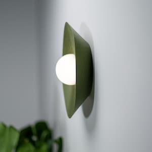 May include: A green wall sconce with a white light bulb. The sconce is made of a textured material and has a curved shape.