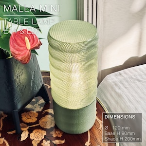 May include: A green table lamp with a woven shade and a cylindrical base. The lamp is 120 mm in diameter, 90 mm tall at the base, and 200 mm tall at the shade. The lamp is labeled "MALLA MINI TABLE LAMP E14 & E27".