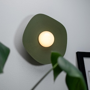 May include: A green wall sconce with a round, white light bulb in the center. The sconce is mounted on a white wall.