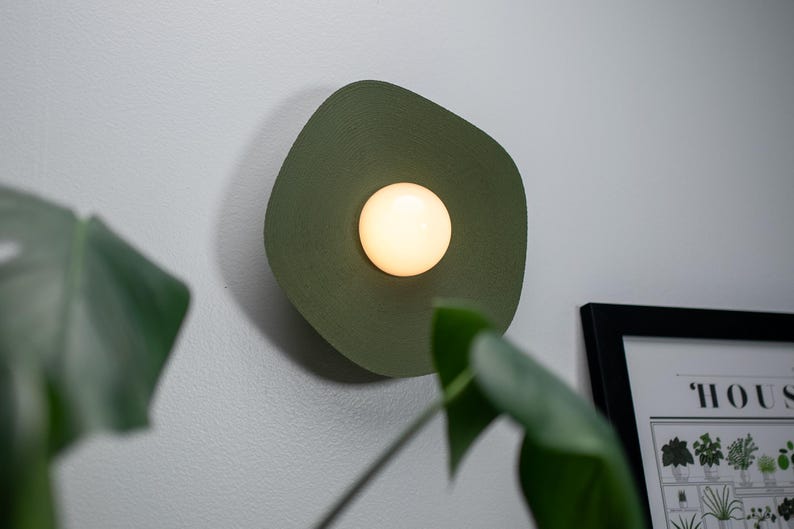 May include: A green wall sconce with a round, white light bulb in the center. The sconce is mounted on a white wall.