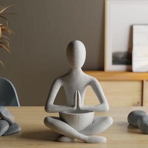 Może przedstawiać: Biała ceramiczna rzeźba przedstawiająca osobę w pozycji siedzącej medytacji z dłońmi złożonymi przed klatką piersiową. Rzeźba ma fakturowaną powierzchnię i trzyma małą miskę.