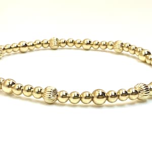 May include: A gold-colored beaded bracelet. The bracelet features a series of small, round beads interspersed with larger, textured beads. The beads are strung together to form a flexible band, suitable for wrist wear.