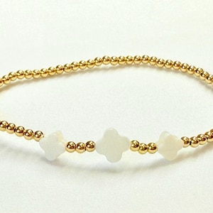 May include: Gold beaded bracelet with three white clover-shaped charms. The bracelet is made of small, round gold beads strung together. The charms are evenly spaced and add a touch of elegance.