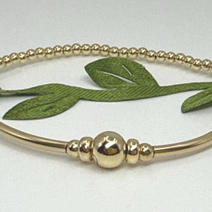May include: Gold-colored beaded bracelet. The bracelet features small, round beads along the top half and a larger central bead with smaller beads on either side. The bracelet is set against a backdrop of green leaves.