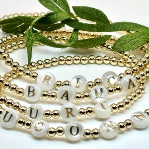 May include: A set of four gold bead bracelets with white beads that have gold letters spelling out the name "BARICIN" in a stacked design.