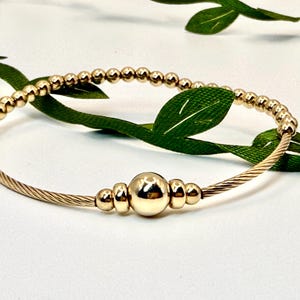 May include: A gold-colored bracelet with a twisted cable design and a series of small, round beads. The bracelet features a larger, central bead and smaller beads. The jewelry is displayed on a white surface with green leaves.