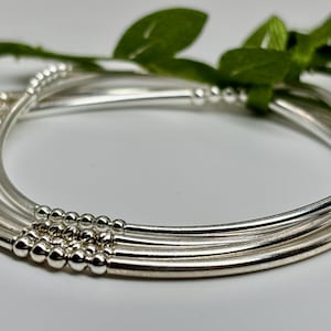 May include: A set of silver-colored bangles with small beaded accents. The bangles are arranged in a curved formation, with green leaves in the background. The jewelry is displayed on a white surface.
