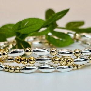 May include: Three stacked bracelets with silver and gold oval beads. The bracelets are made of metal and have a polished finish.