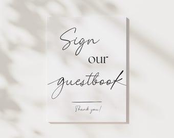 May include: A white sign with black script that reads "Sign our guestbook" with "Thank you!" at the bottom. The sign is rectangular and has a minimalist design, suitable for weddings or events.