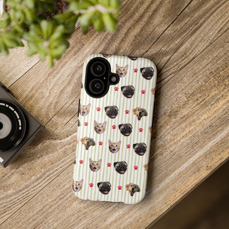 Puede incluir: Funda de tel&eacute;fono blanca con un patr&oacute;n repetido de caras de perros y gatos, junto con huellas de patas rojas, sobre un fondo de rayas verticales. La funda tiene un recorte para una c&aacute;mara de doble lente.