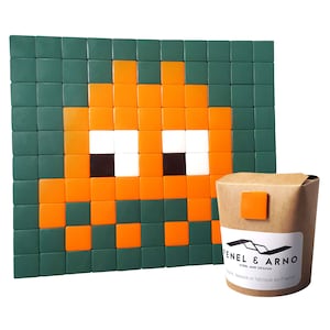 May include: A green and orange pixel art design of a space invader. The design is made up of small square tiles. The image also includes a brown paper box with the text "Fenel & Arno Steel and Design" and "Conçu, dessiné et fabriqué en France".