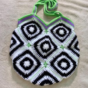 May include: A handmade crochet tote bag with a geometric pattern. The bag features a repeating pattern of black and white squares, accented with green and purple trim. The handles are a bright green color.