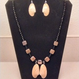 May include: A necklace and earring set displayed on a black stand. The necklace features a black beaded chain, brown square beads, and two large, beige shell pendants. The earrings have matching shell pendants.