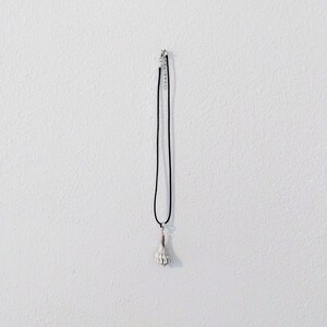 May include: A necklace featuring a thin black cord and a small, off-white seashell pendant. The pendant is attached to the cord with a silver-toned metal bail, and the necklace includes a silver-toned metal chain extender with a lobster claw clasp.