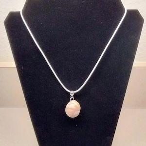 May include: A light beige pendant necklace with a round, textured stone. The pendant hangs from a thin, white cord. The necklace is displayed on a black jewelry stand.