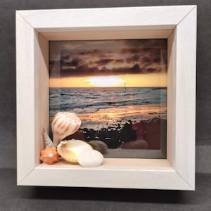 May include: A framed beach scene with a sunset over the ocean. The frame is white, and seashells are placed in front of the image. The shells include a large, striped conch shell, a small orange shell, and a white, flat shell.