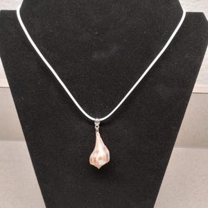 May include: A seashell pendant necklace. The pendant is a light brown and white seashell suspended from a white cord. The necklace is displayed on a black jewelry stand.