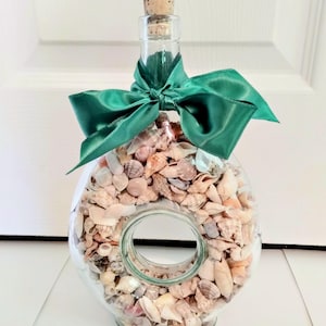 May include: A clear glass donut-shaped bottle filled with various small beige, tan, and white seashells. The bottle features a cork stopper at the top and is decorated with a wide, dark green satin ribbon tied into a bow around the bottle neck.
