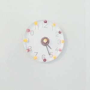 May include: A round white wall clock featuring a dotted numeral design from 1 to 12. The clock face is decorated with small, alternating yellow and pink spherical accents placed at each hour mark. A central circular element holds two gray hands.