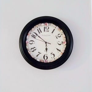 May include: A round wall clock with a black frame and a white face featuring black Arabic numerals. The inner border of the clock face is decorated with small, light-colored seashells. The brand name Sterling & Noble is printed in the center.