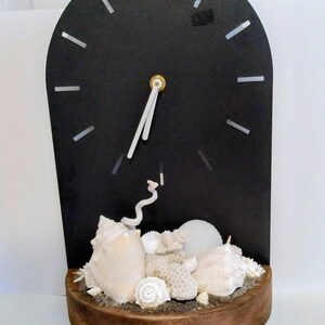 May include: An arched black wall clock featuring silver hour markers and white hands. The base is a rounded wooden platform decorated with an assortment of white seashells, coral fragments, and sand, creating a textured, coastal-inspired display.