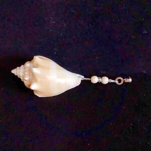 May include: A cream-colored spiral seashell charm featuring a pointed apex and a wide opening. The shell is attached to a thin cord adorned with two small, round, off-white beads and a silver-toned metal jump ring at the end for attachment.