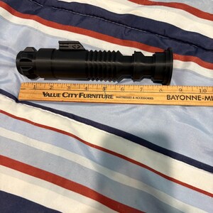 May include: A black plastic lightsaber prop with a ribbed design and a silver accent. The prop is approximately 9 inches long.