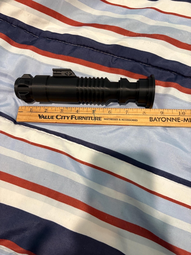 May include: A black plastic lightsaber prop with a ribbed design and a silver accent. The prop is approximately 9 inches long.