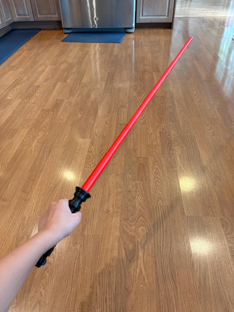 May include: A red lightsaber toy with a black handle. The lightsaber is held by a hand in a wooden room.