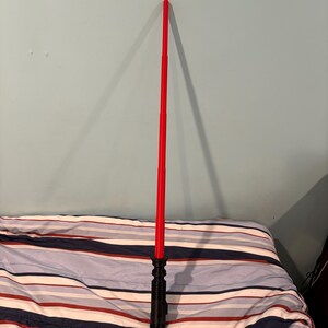May include: A red lightsaber toy with a black handle. The lightsaber is extended and standing upright on a striped bedspread.