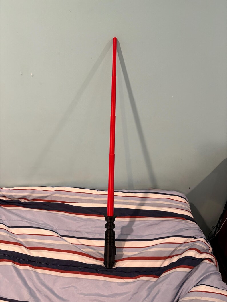 May include: A red lightsaber toy with a black handle. The lightsaber is extended and standing upright on a striped bedspread.