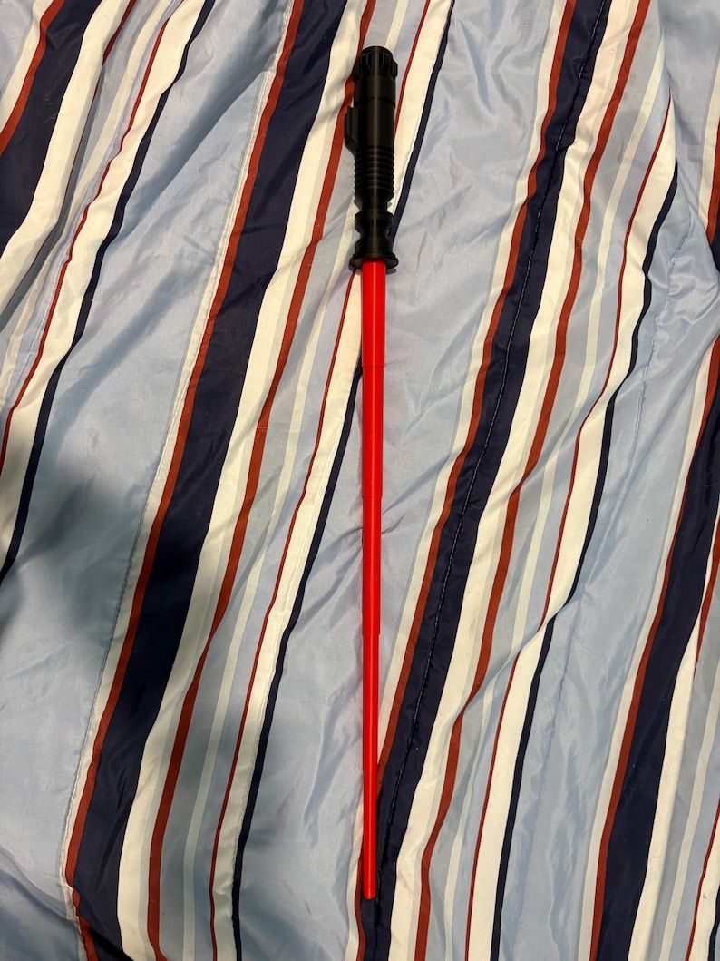 May include: A red lightsaber toy with a black handle. The lightsaber is lying on a blue and white striped fabric.