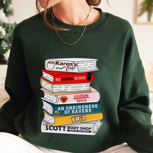 May include: Dark green sweatshirt featuring a stack of illustrated books. The books have titles like "Karen's Cafe," "Red Bedroom Records," and "An Unkindness of Ravens." The bottom book reads "Scott Body Shop, Service and Repair."