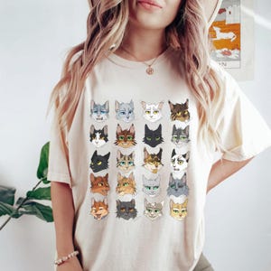 May include: A cream-coloured t-shirt featuring a graphic print of 24 cat faces arranged in a grid pattern. The cats are depicted in a cartoon style with various expressions and fur colours.