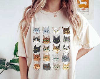 May include: A cream-colored t-shirt with a graphic print of 24 cat faces in a grid pattern. The cats are drawn in a cartoon style with different expressions and fur colors.