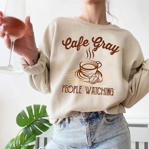 May include: A beige sweatshirt with the text "Cafe Gray" and a stylized illustration of a coffee cup with steam rising from it. The text "People Watching" is below the illustration.