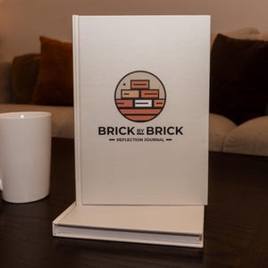 以下が含まれることがあります： 「Brick by Brick - Reflection Journal」というタイトルと、円の中に積まれたレンガのロゴが描かれた白いハードカバーのジャーナルです。