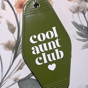 May include: Olive green keychain with the words "cool aunt club" in white, along with a white heart symbol. The keychain has a retro motel key design and a silver key ring.