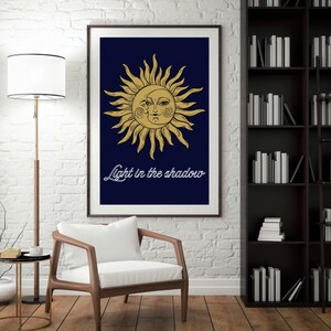 Può includere: Stampa artistica incorniciata con un'illustrazione dorata del sole e della luna su sfondo blu scuro. Il testo "Light in the shadow" è scritto in bianco. L'opera d'arte è esposta in una stanza con un muro di mattoni bianchi.