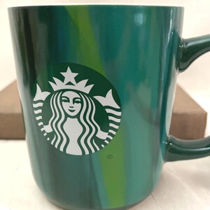 May include: A glossy ceramic mug featuring a swirling pattern of dark and light green hues. The front displays the white Starbucks siren logo, which includes a crowned figure with flowing hair. The mug has a curved handle on the right side.