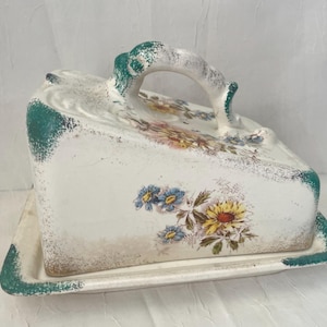 May include: A white ceramic butter dish with a lid and handle, featuring floral designs in yellow, blue, and pink. The edges and handle are accented with green paint, and the dish has a rectangular base.