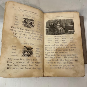 Puede incluir: Libro abierto con páginas envejecidas y amarillentas con texto e ilustraciones. La página izquierda tiene texto sobre trineos y un nido de pájaros. La página derecha tiene texto sobre la luna y las estrellas. El libro se titula "SANDERS' UNION READER."