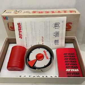 May include: A vintage board game, "Jitters," with a red timer, cards, and instructions. The game box is open, revealing the components inside. The game's name is printed on the box and components.