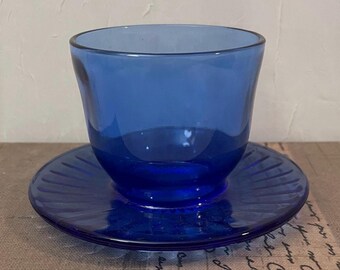 May include: A cobalt blue glass teacup and saucer set. The teacup has a rounded shape and sits on a matching saucer with a ridged design. The set is made of transparent glass, reflecting light and creating a classic aesthetic.