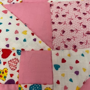 Puede incluir: Primer plano de un edredón de patchwork con temática de corazón. El edredón presenta varias telas rosas y telas blancas con patrones de corazón en rojo, amarillo y azul. El edredón está hecho de cuadrados y triángulos.