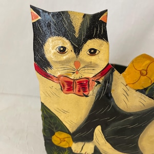 May include: A decorative metal container shaped like a cat with black and cream fur, wearing a red collar with a bow. The cat is depicted sitting in green grass with yellow flowers. The metal surface features painted details and a glossy finish.