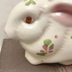 May include: A white ceramic rabbit figurine featuring a hand-painted floral pattern with small pink flowers, green leaves, and yellow centers. The rabbit has a glossy finish, a brown eye, and soft pink shading on the interior of its long ear.